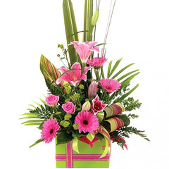 A vibrant pink and green box of lilies, roses, mini gerberas, and chrysanthemums with tropical foliage and spear grass.